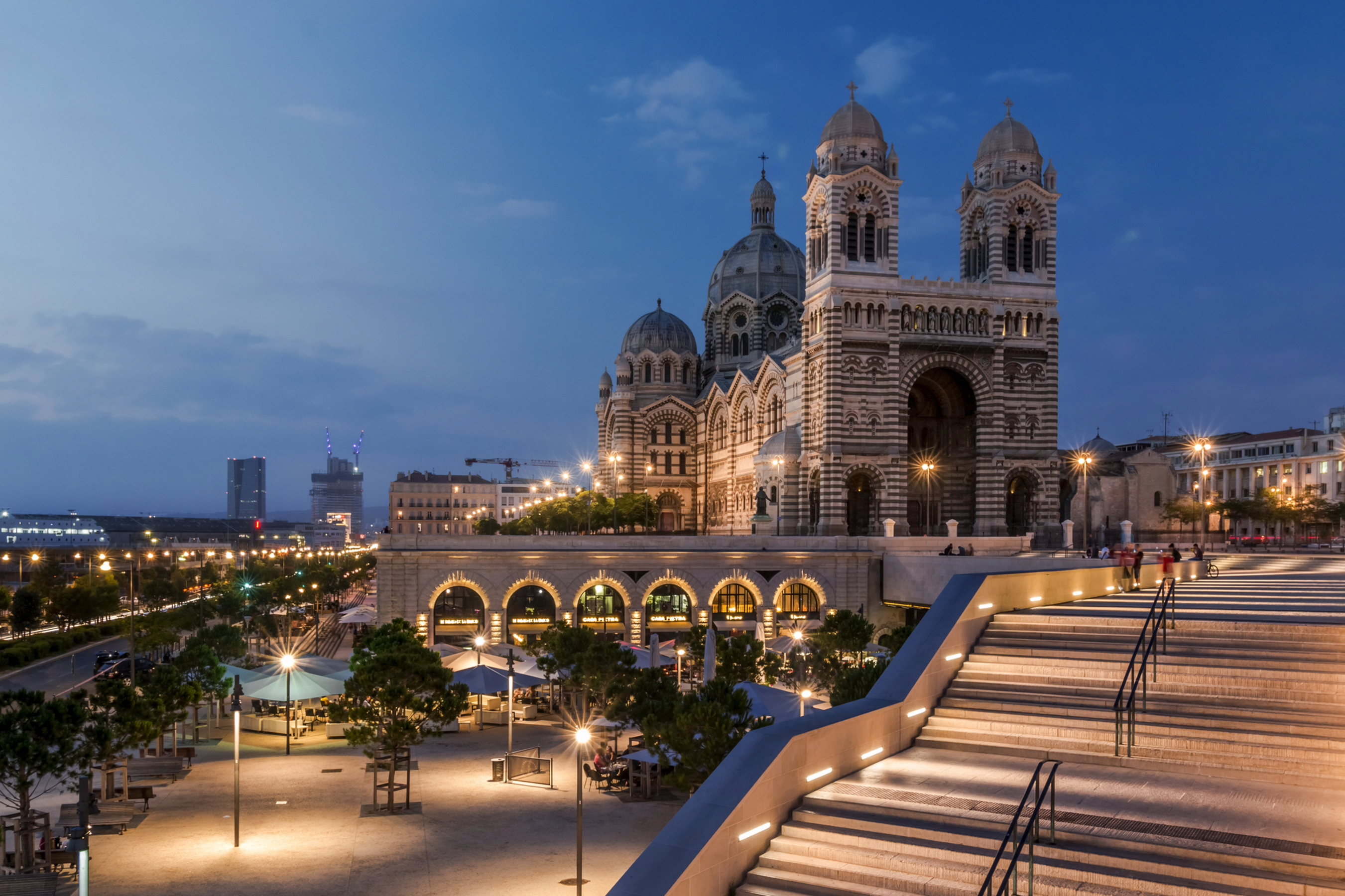 Basilika bei Dämmerung mit beleuchtetem Vorplatz und Treppe in urbaner Umgebung.